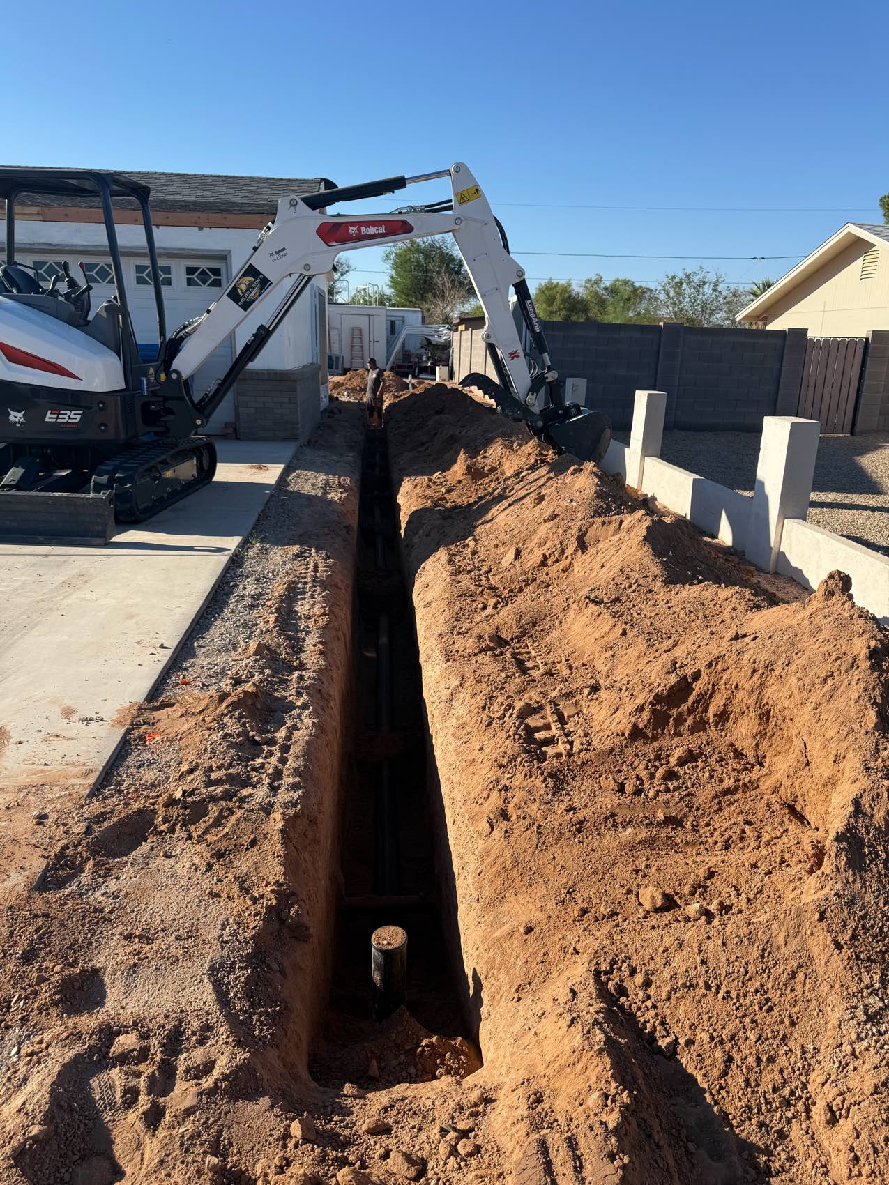 Open trench for a sewer main running the length of a residential lot, with a Bobcat E35 excavator parked on the driveway.
